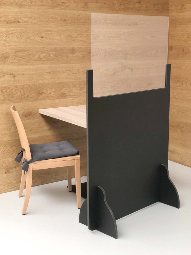 Das Foto zeigt einen Sitzplatztrenner in schwarz und einem milchigen Glasfenster vor einem Tisch und einem Stuhl stehend.