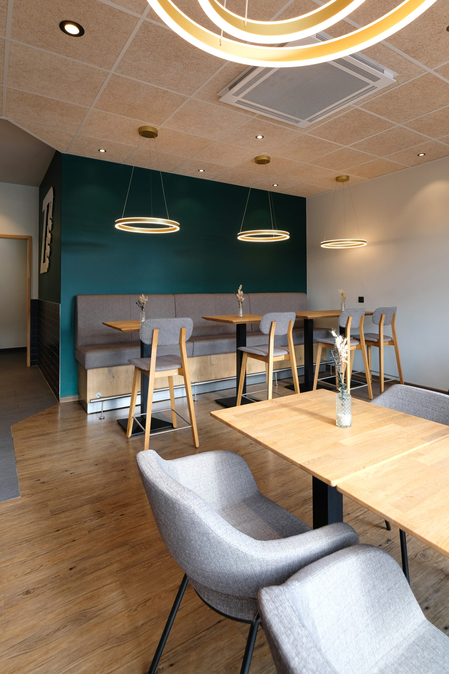 Moderner Sitzbereich mit gepolsterten Hochlehnbänken und hellgrauen Holzstühlen, vor einer smaragdgrünen Wand und unter drei schwebenden LED-Ringlampen – im Vordergrund ein heller Holztisch mit zwei bequemen Sesseln.