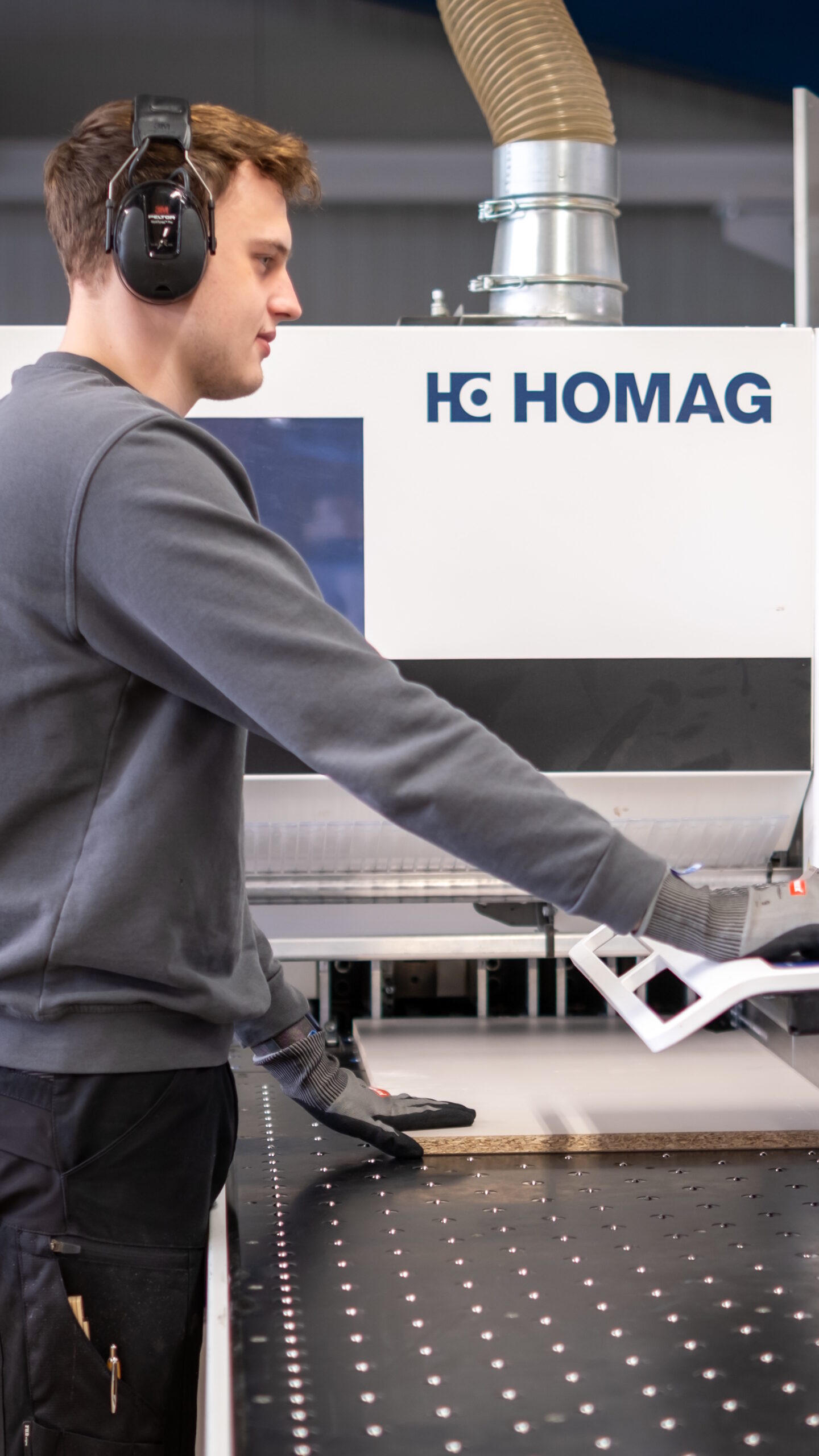 Mitarbeiter mit Gehörschutz und Handschuhen bedient eine HOMAG-Maschine zur Holzbearbeitung in einer Werkhalle.