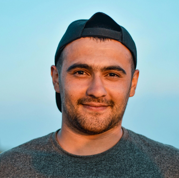 Porträt eines jungen Mannes mit dunkler Baseballkappe, der leicht lächelt und ein graues T-Shirt trägt, vor hellblauem Himmel im Hintergrund.