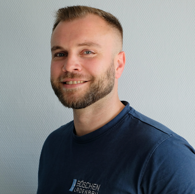 Porträt von Prokurist Stefan Kreklau in dunkelblauem T-Shirt mit Bö­schen Ladenbau-Logo, vor hellem strukturiertem Hintergrund.