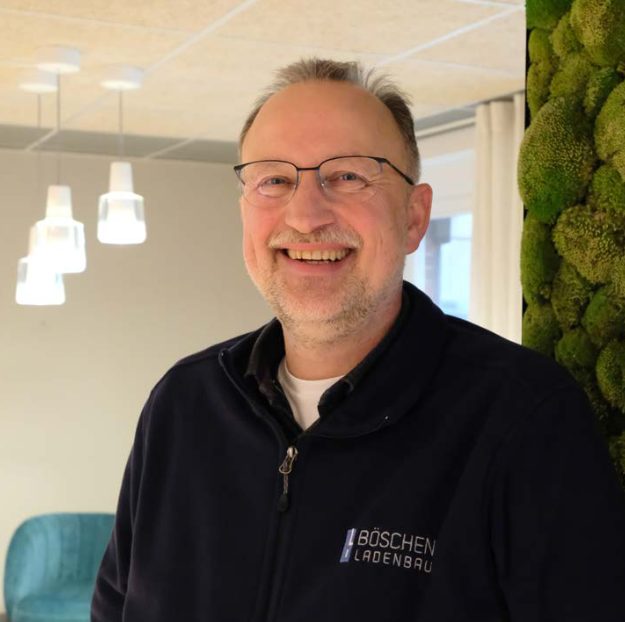 Ein lächelnder Mann in Arbeitskleidung von „Böschen Ladenbau“ steht in einem modern gestalteten Innenraum.