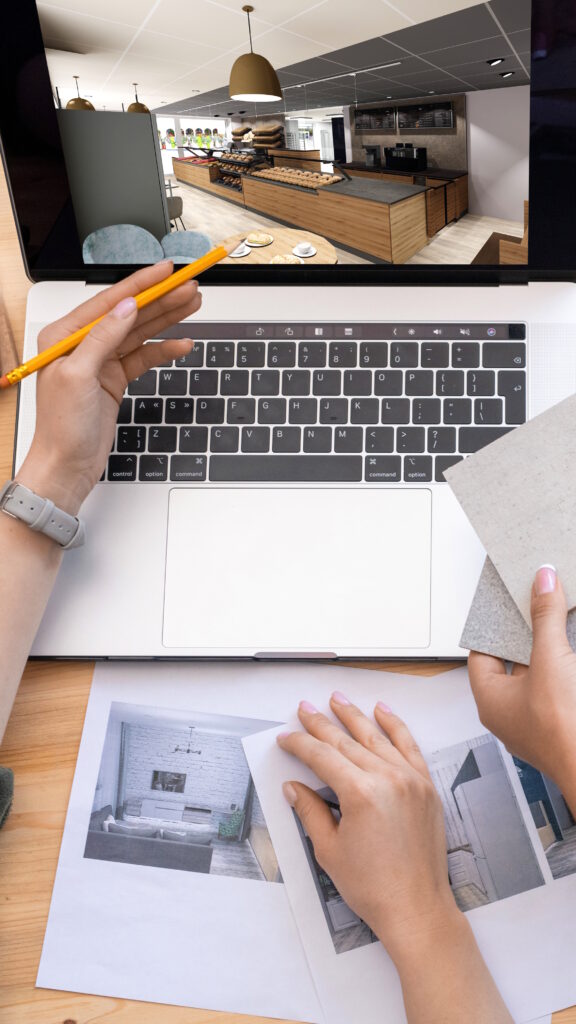 Zwei Mitarbeiter planen eine Innenraumgestaltung am Laptop mit 3D-Visualisierung eines Bäckereibereichs, Musterplatten und ausgedruckten Entwürfen auf dem Tisch.