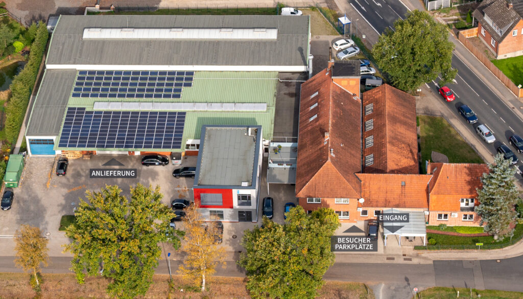 Luftaufnahme der Firma Böschen ladenbau GmbH in Ottersberg mit Produktionshalle, Solarzellen auf dem Dach, Bürogebäude und angrenzender Straße. Ebenso gibt es eine Beschriftung wo der Eingang, die Besucherparkplätze und die Anlieferung ist.