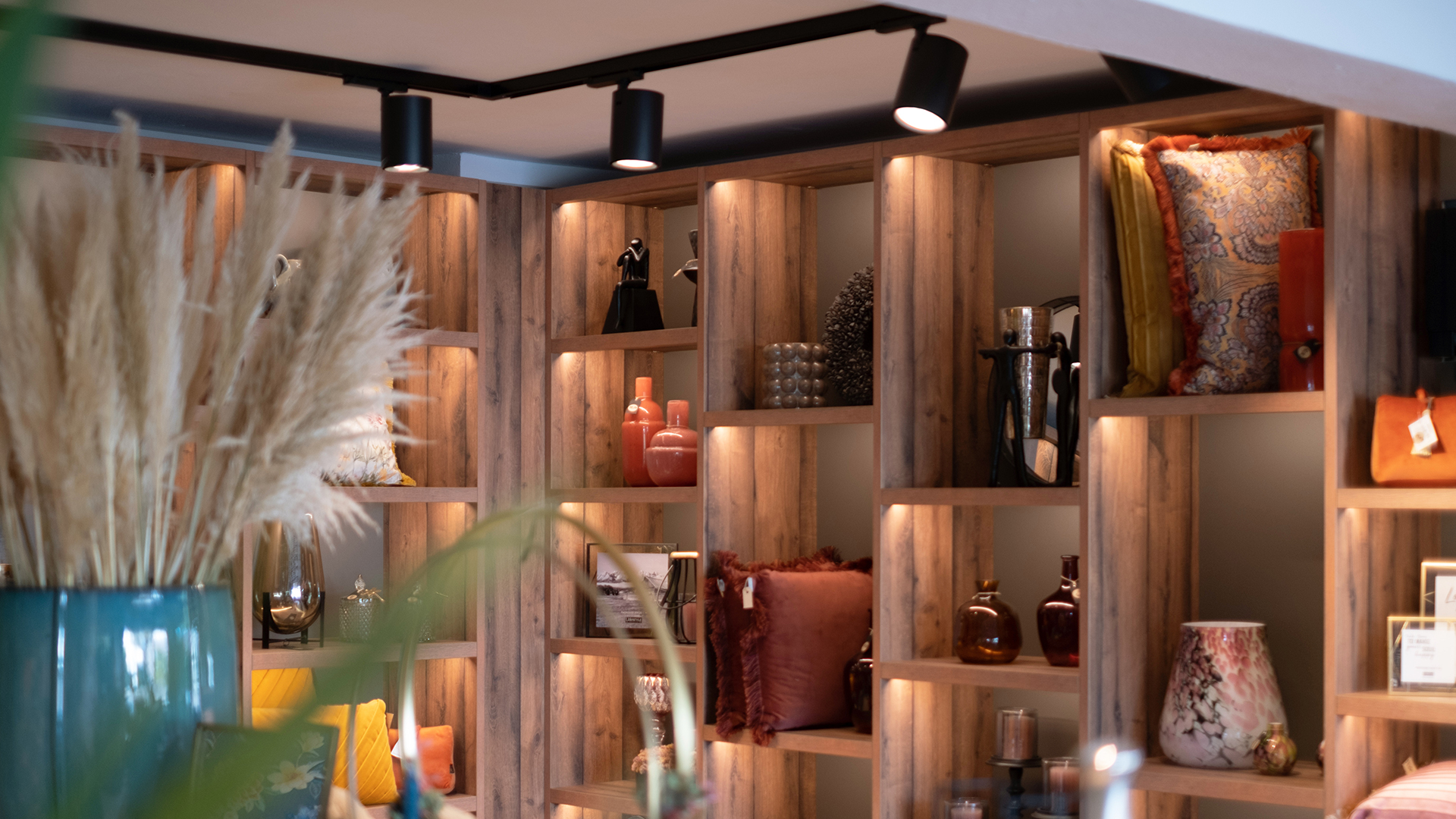 Nahaufnahme eines beleuchteten Regalsystems in Holzoptik mit schwarzen Strahlern die auf verschiedene Produkte eines Dekoladens leuchten u.a. Figuren, Vasen, Kissen.