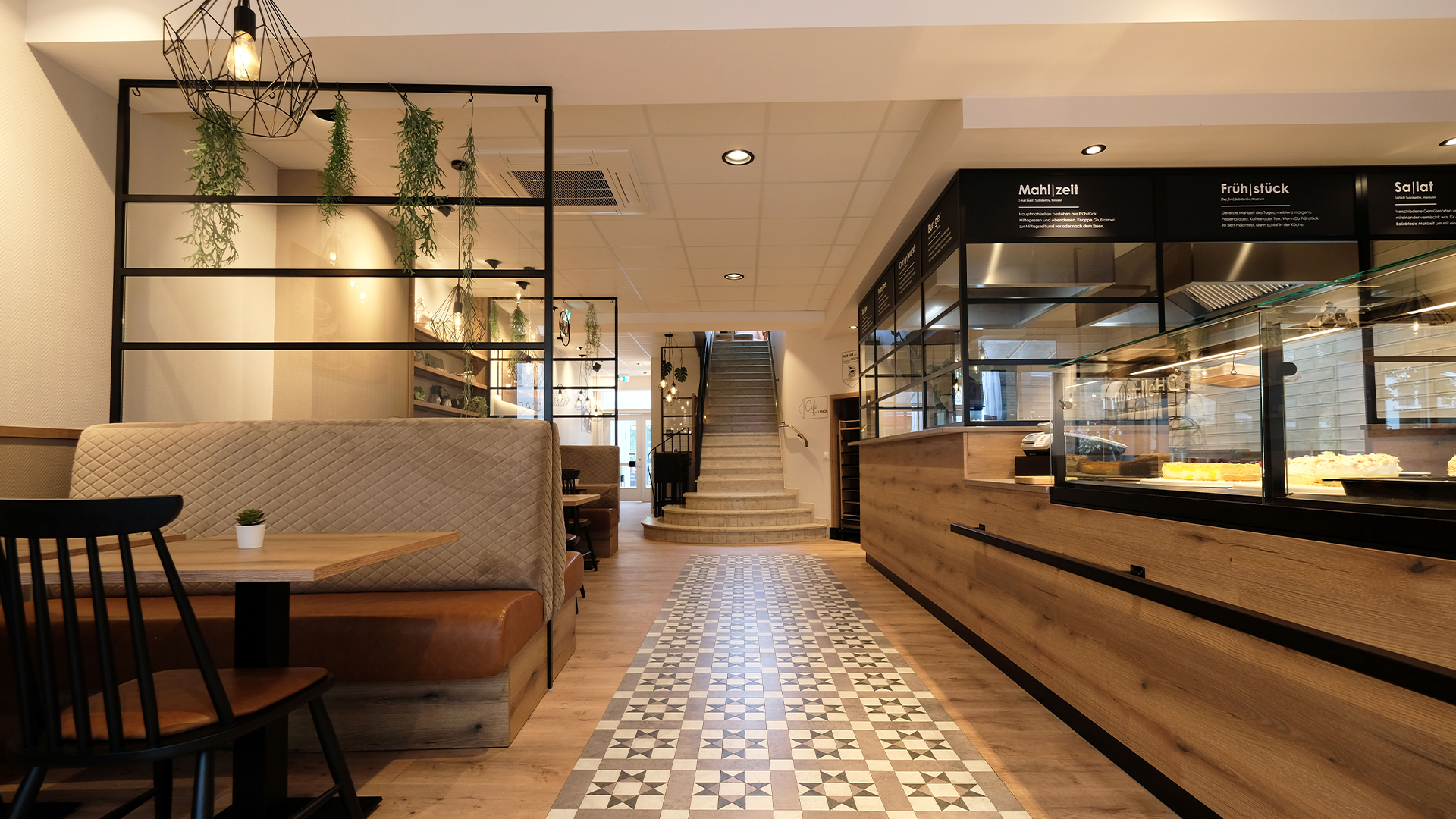 Ein mittiger Blick auf den Eingang eines Café-Restaurants. Auf der rechten Seite ist der Tresen in Holzoptik und mit schwarzen Metallelementen zu sehen. Links der Sitzbereich mit schönen gemütlichen Sitzbänken und Stühlen in braun und beige Stühlen sowie Metallelementen die als Raumtrenner dienen und zur Deko mit Pflanzen beschmückt sind.
