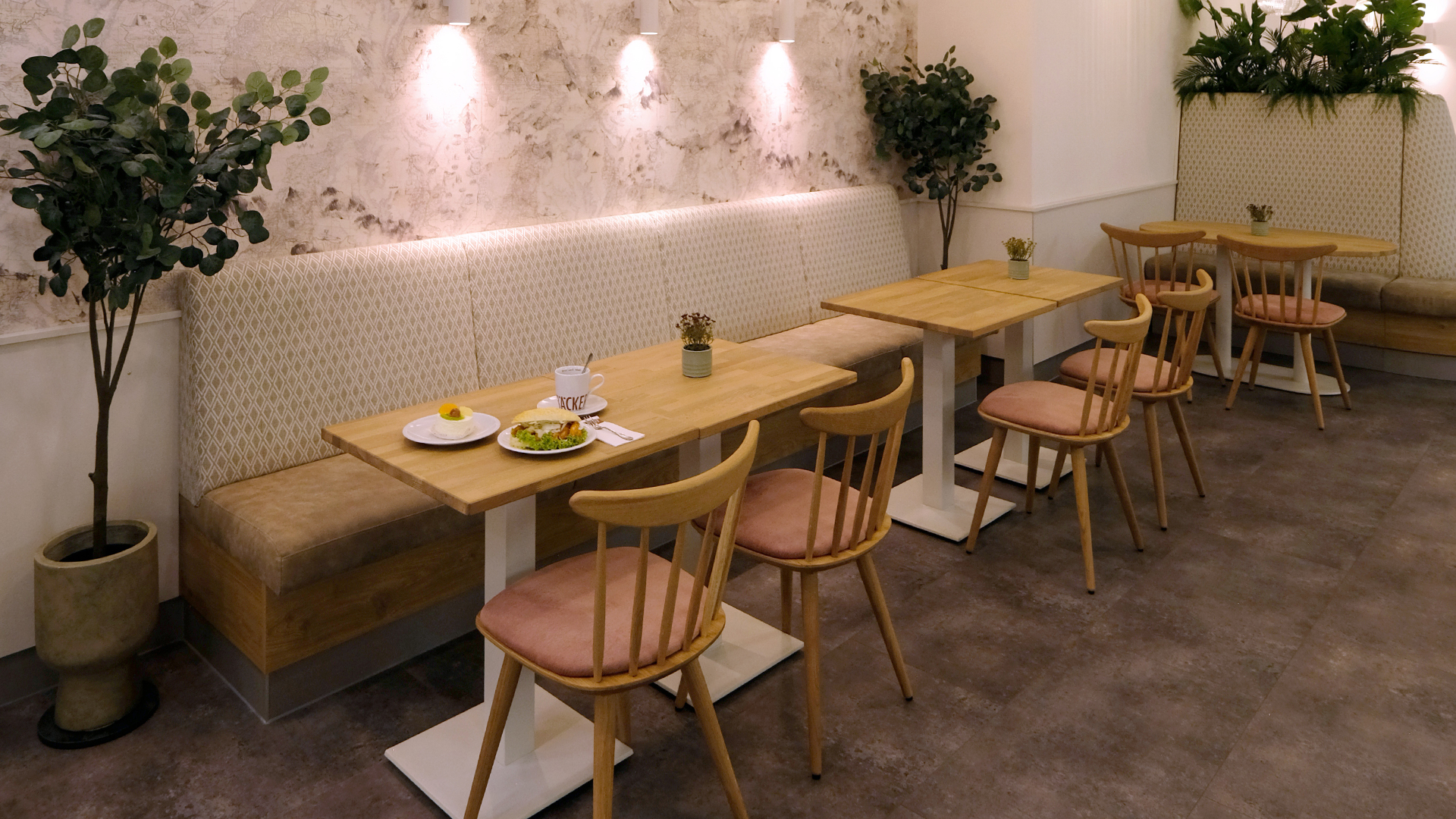 Moderner heller Sitzbereich in hellen weiß, beige, braun und Rosetönen mit einer tollen Wandtapete und weißen Beleuchtungsstrahlern im Hintergrund und einem leckerem Frühstück auf dem Tisch. Die Tischgestelle bestehen aus einer Holzplatte und einem weißen Fußgestell aus Metall. Im Hintergrund ist noch eine maßgefertigte Sitzbank zu sehen in der im oberen bereich Pflanzen eingearbeitet wurden, welche direkt als Sichtschutz dienen. Dekoriert ist der Sitzbereich mit grünen Pflanzenelementen und Kunstbäumen.