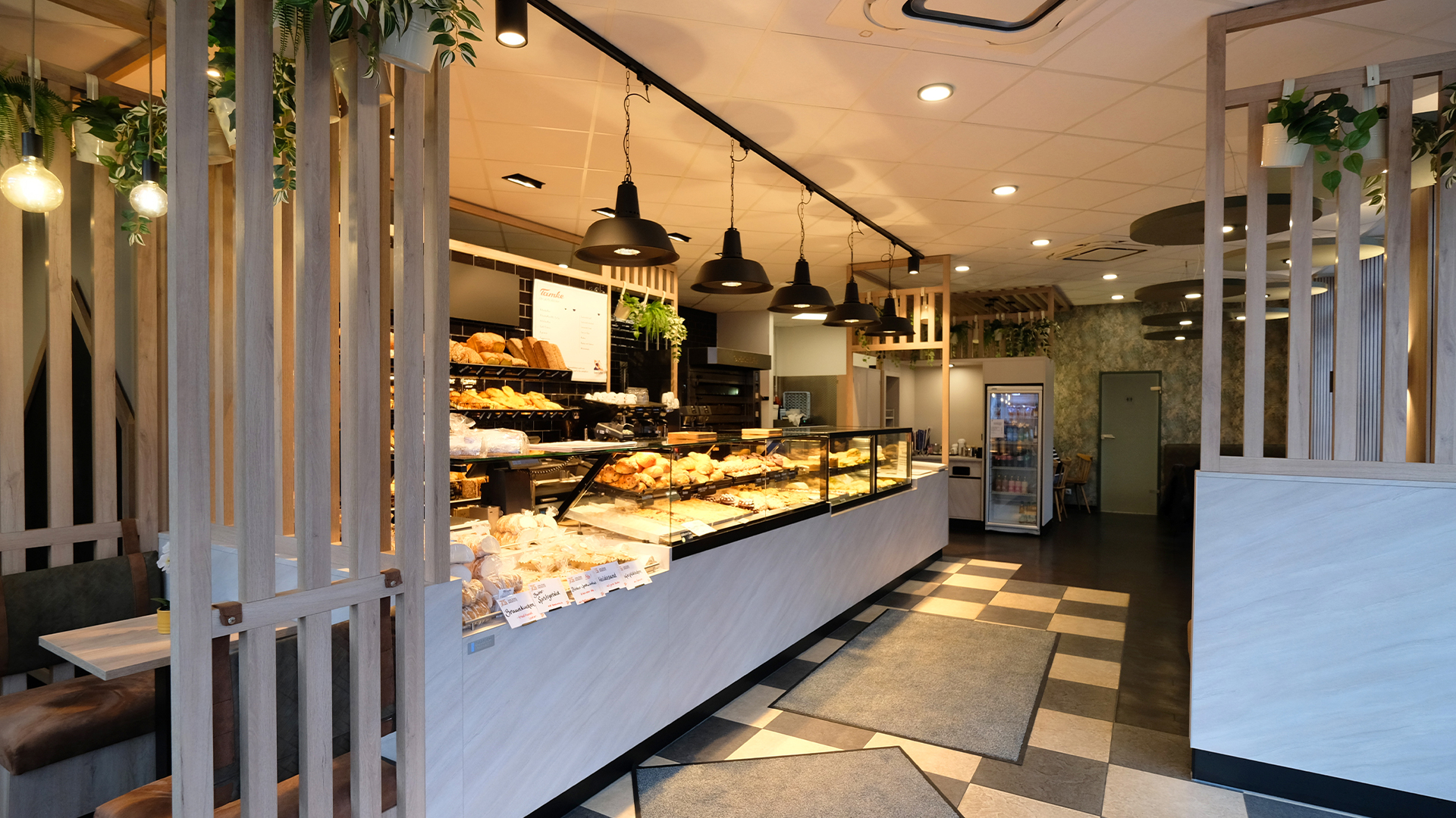 Ansicht weißer heller schöner Bäckereitresen mit schwarzen Hängeleuchten und Strahlern versehen. Volles übersichtliches Brotregal mit einem weiß-schwarzem Kachelboden und Lamellen-Trennelementen für den Sitzbereich.